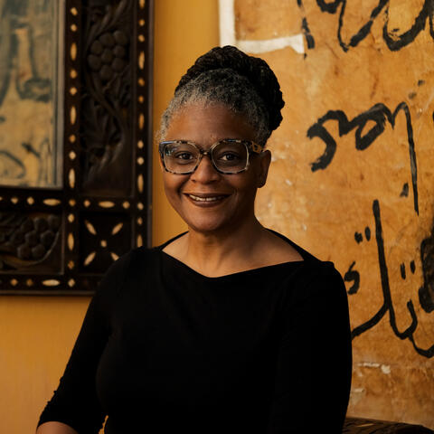 Photo of woman smiling, dressed in black with a gold painted wall behind her.